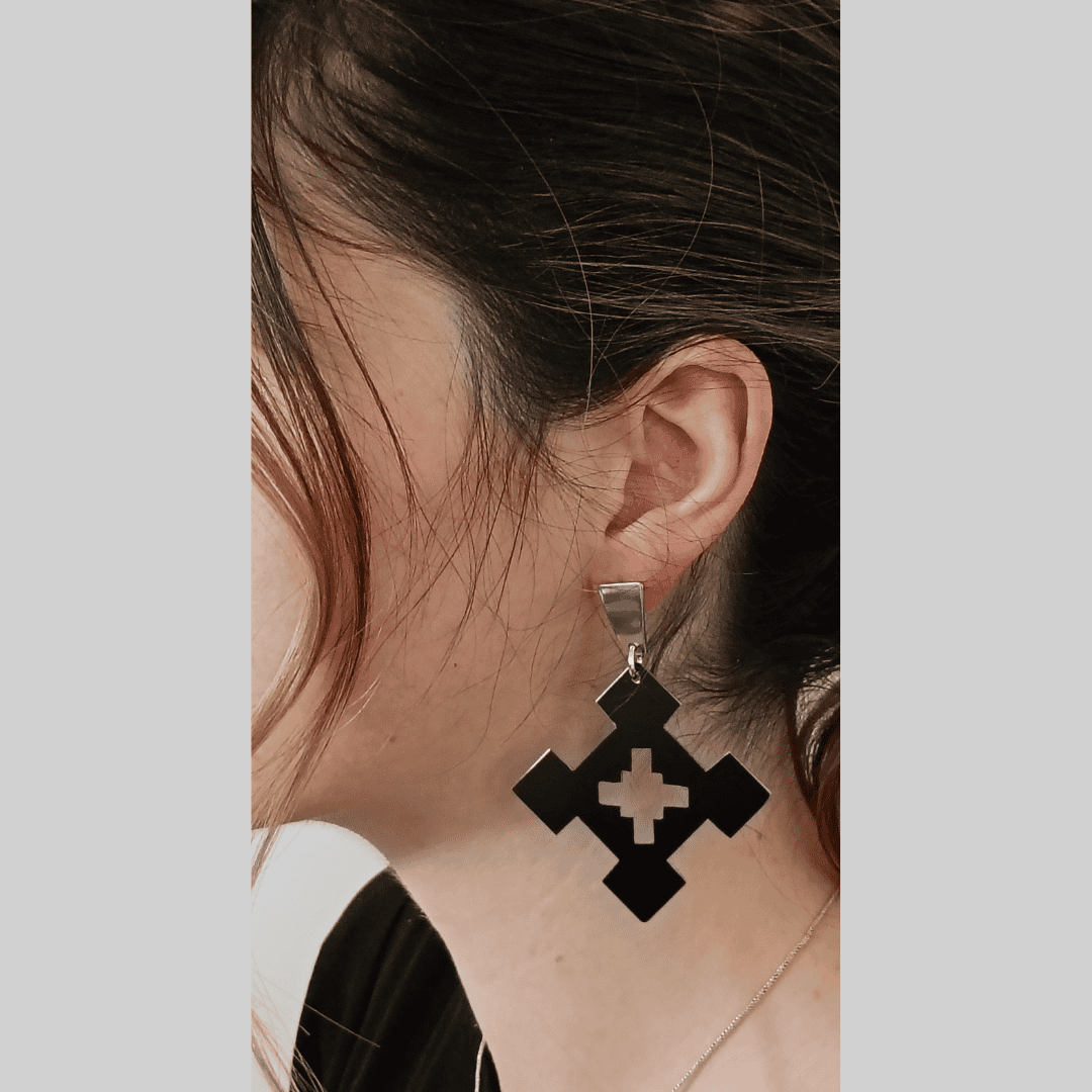 Close-up shot of a person's ear adorned with a pair of eye-catching earrings. The earrings feature a unique design: a black square shape with a cut-out cross pattern in the center. The square is suspended from a sleek silver hook, providing a beautiful contrast. The overall style is minimalist yet modern, with a touch of edgy sophistication. The mood is chic and contemporary, reflecting the modern design of the jewelry.