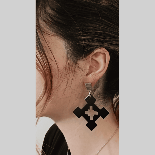Close-up shot of a person's ear adorned with a pair of eye-catching earrings. The earrings feature a unique design: a black square shape with a cut-out cross pattern in the center. The square is suspended from a sleek silver hook, providing a beautiful contrast. The overall style is minimalist yet modern, with a touch of edgy sophistication. The mood is chic and contemporary, reflecting the modern design of the jewelry.