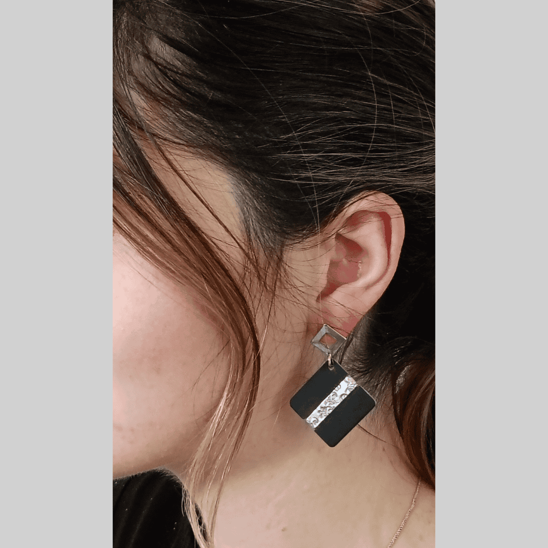 Close-up view of a woman's ear adorned with a pair of stylish earrings. The earrings are square-shaped with a black base and a silver vertical stripe featuring a textured or sparkling detail. The silver detailing contrasts sharply with the matte black, creating a modern and minimalist aesthetic. The earrings hang from simple silver hooks, adding to the overall clean lines and geometric design. The woman's dark brown hair frames her face, with a few strands gently falling around her ear.