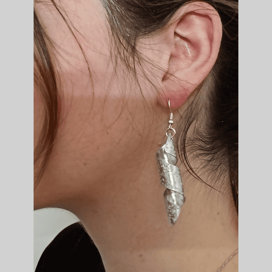 Close-up of a person's ear wearing a pair of long, silver dangle earrings. The earrings are uniquely designed in a spiral shape, with a textured, aluminum surface. The texture appears to be a stamped pattern. The earrings have a modern and contemporary style.