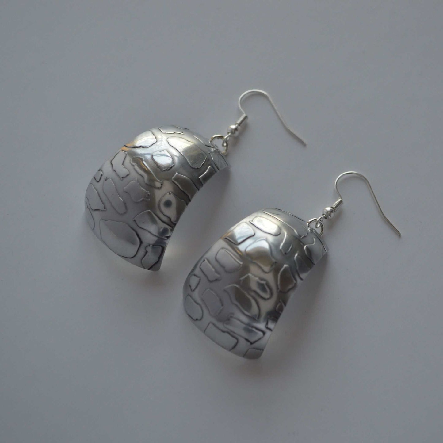 A pair of silver-colored aluminum earrings with a curved and patterned design.