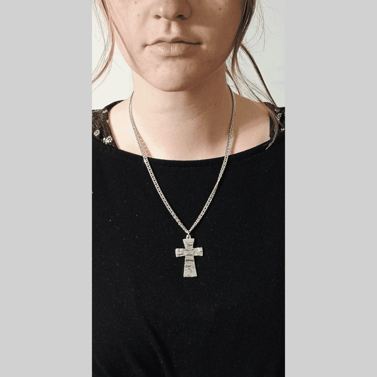 Close-up image showcasing a silver cross necklace worn by a person. The necklace features a textured, silver cross pendant hanging from a cuban link chain. The chain is a medium-weight type and sits comfortably around the neck. The individual is wearing a simple black shirt that provides a neutral background, highlighting the necklace. 