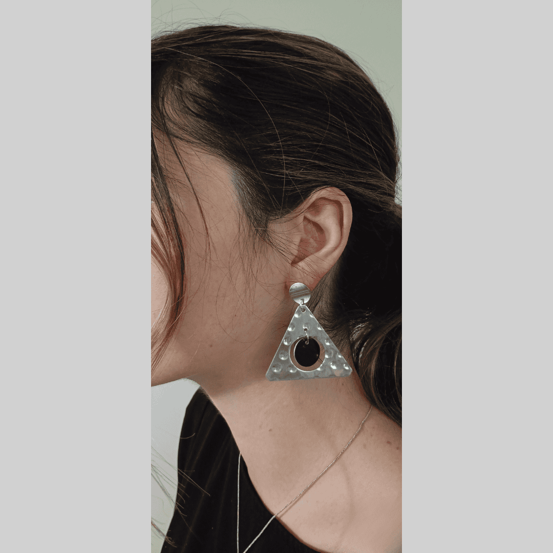 Close-up profile view of a woman wearing a pair of eye-catching silver earrings. The earrings are triangular in shape with a hammered or textured metal surface.  A black circle is centrally located within each triangle. The overall style is minimalist yet bold, with a modern and geometric aesthetic. The background is a muted, neutral color, allowing the silver earrings to stand out. The image has a clean, simple feel, focusing the attention solely on the earrings and their unique design.