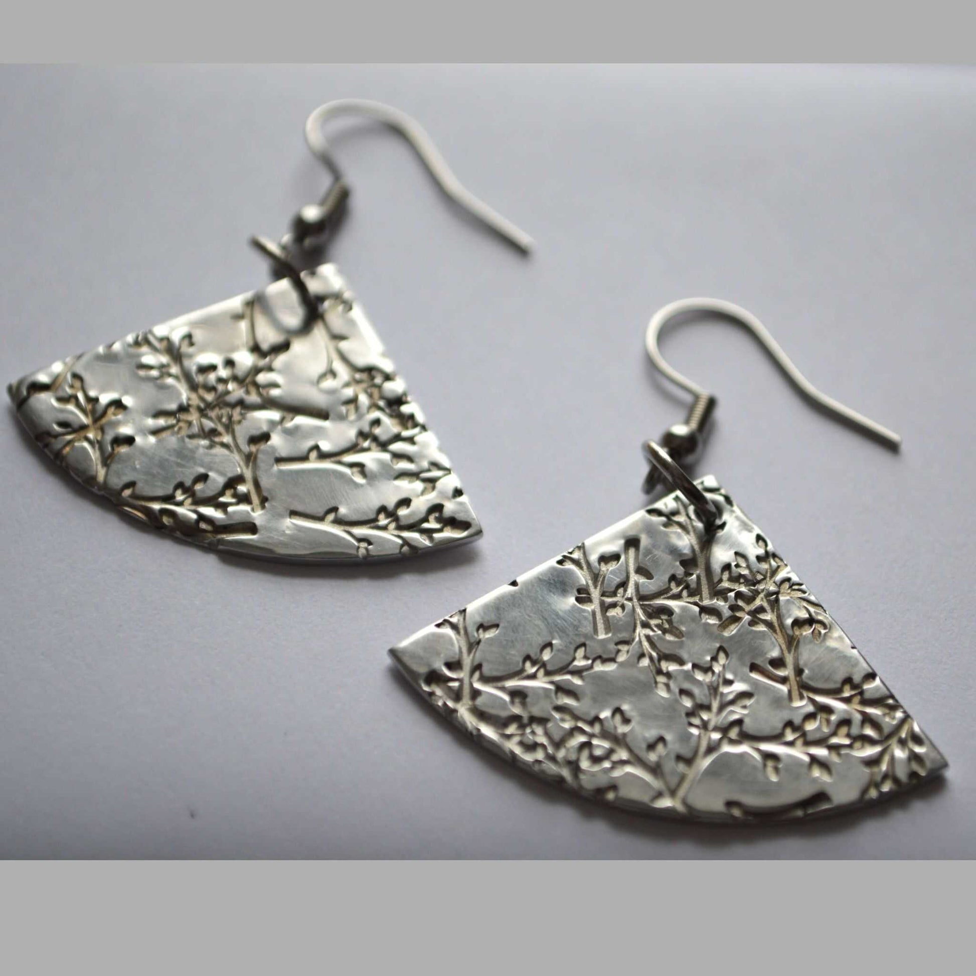 A pair of fan-shaped silver earrings with a stamped floral pattern on a white background.