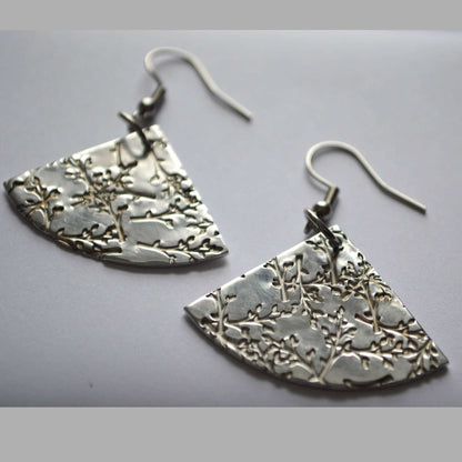 A pair of fan-shaped silver earrings with a stamped floral pattern on a white background.