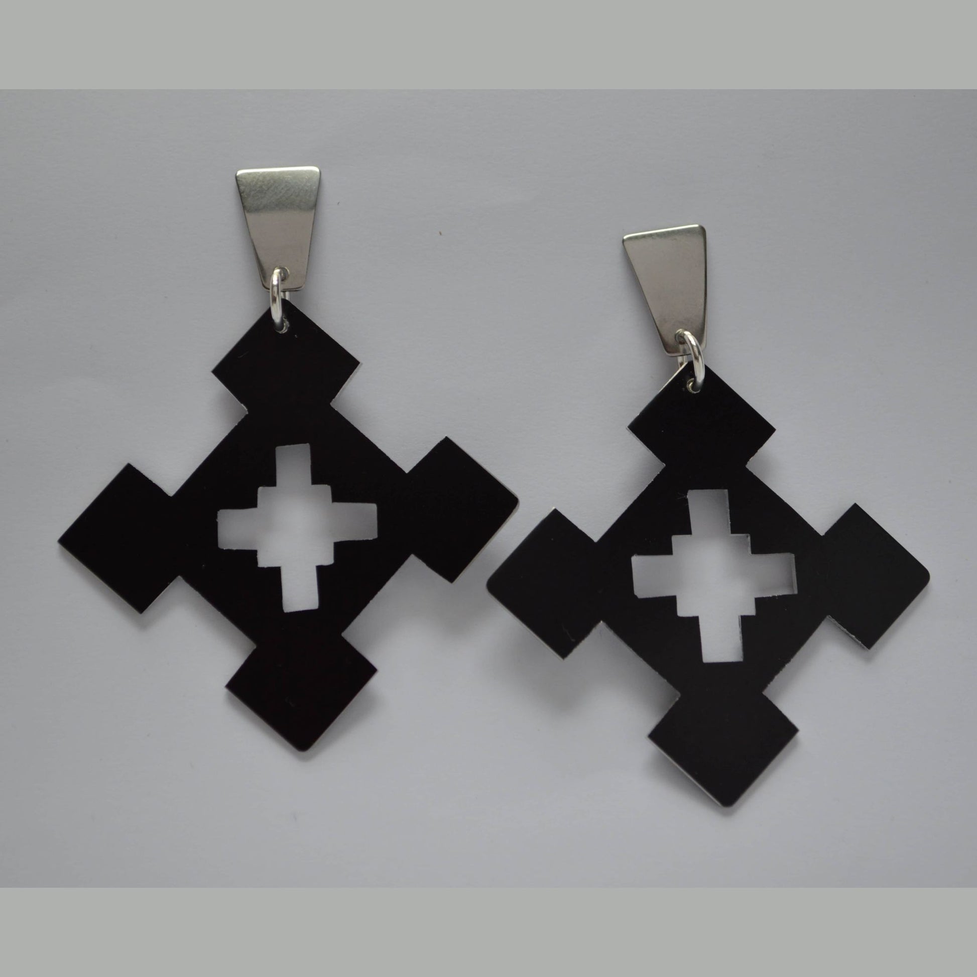 A pair of bold statement earrings with a geometric design, featuring black anodized aluminum diamond shapes and cross cutouts, suspended from polished stainless steel posts.