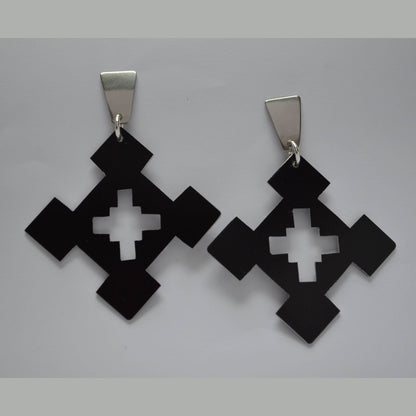 A pair of bold statement earrings with a geometric design, featuring black anodized aluminum diamond shapes and cross cutouts, suspended from polished stainless steel posts.