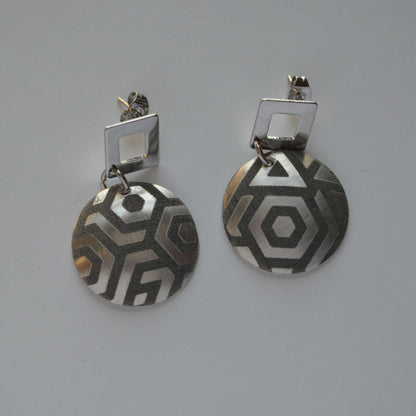 A pair of silver circular earrings with a triangular pattern, attached to a square post.