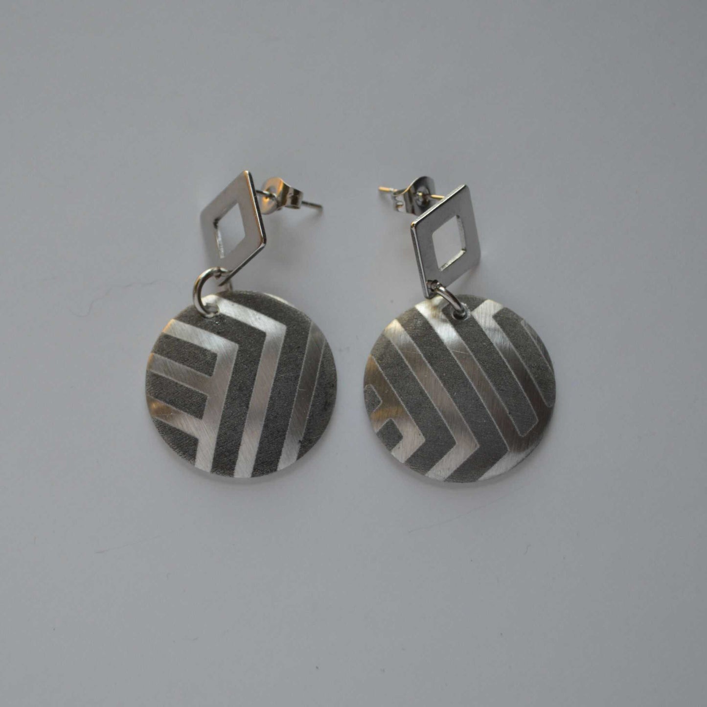 A pair of silver-colored disc earrings with geometric patterns, attached to the ear with a small square post.