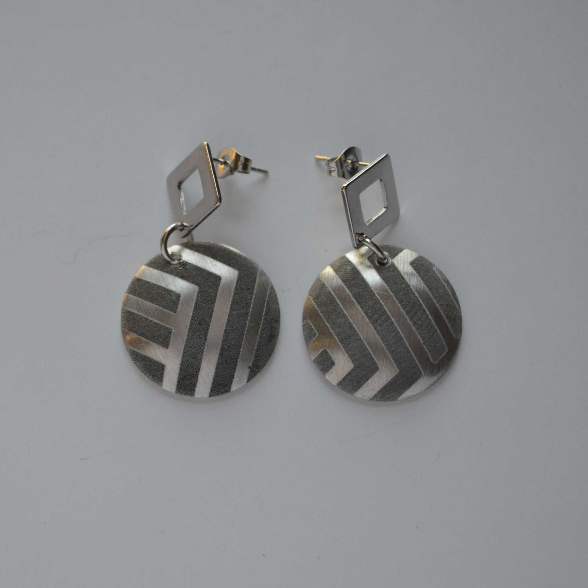 A pair of silver-colored disc earrings with geometric patterns, attached to the ear with a small square post.