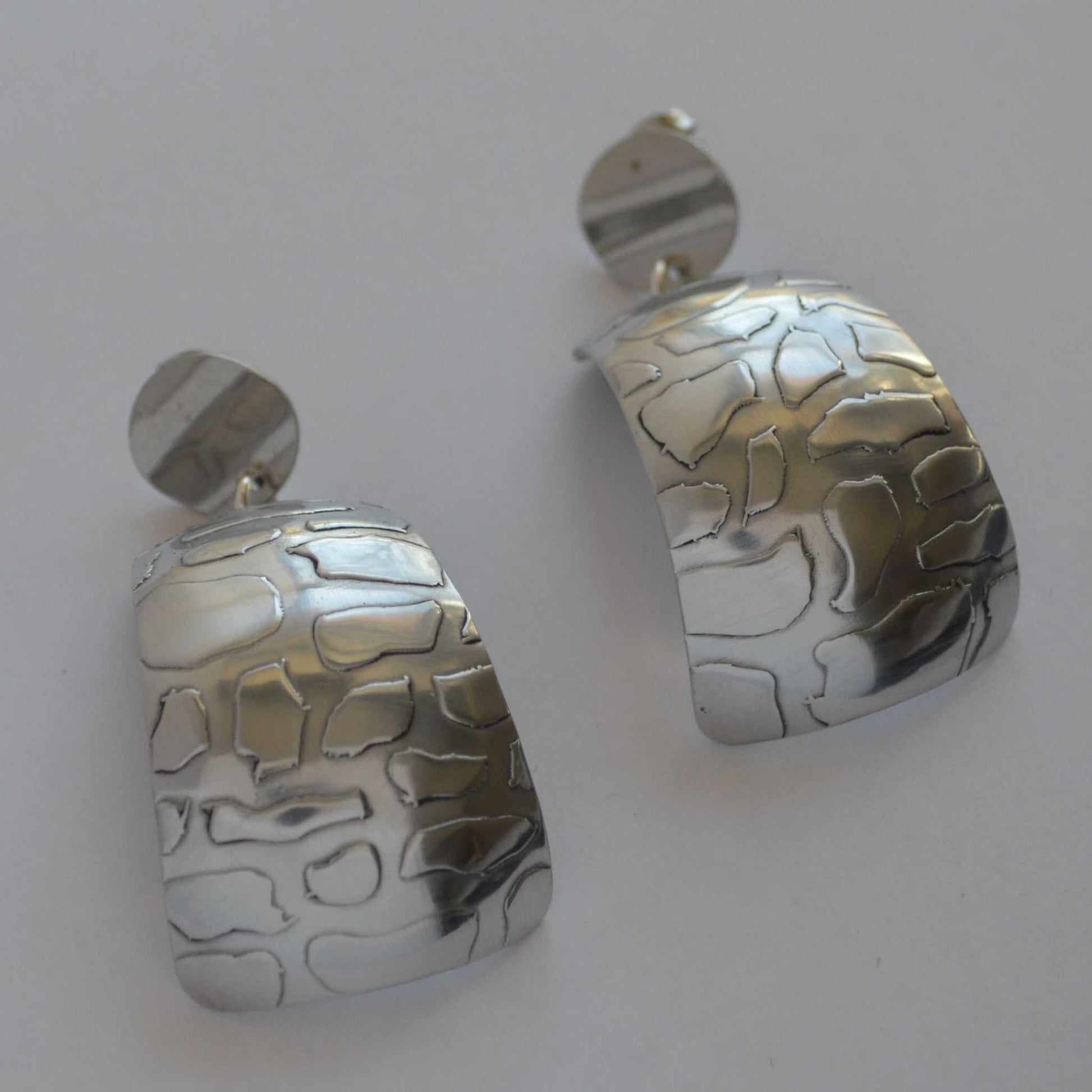 A pair of silver-colored aluminum earrings with a curved and patterned design.