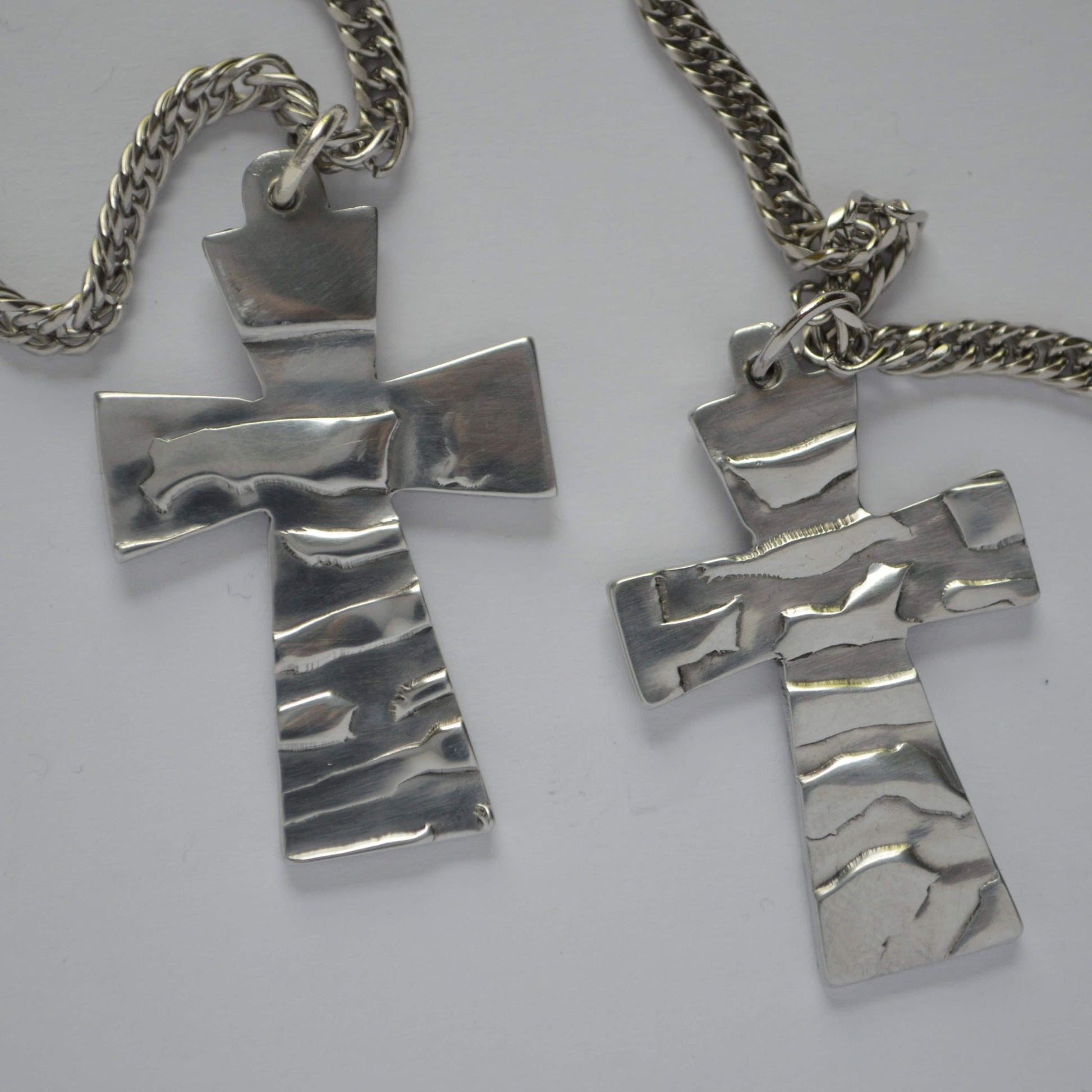 Two silver aluminum cross pendants with a hammered texture and a flowing wave pattern, displayed on a white background.
